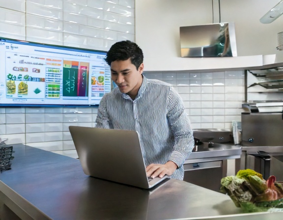 An operations manager working on a laptop in a modern kitchen with a data dashboard on the wall